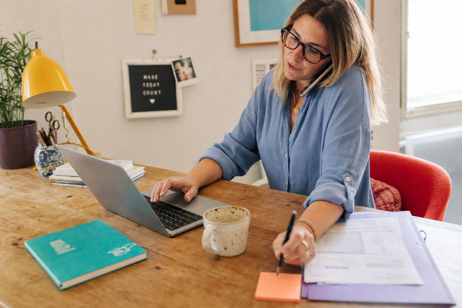 Vrouw met bril werkt thuis aan een laptop, telefoneert en maakt aantekeningen aan een houten bureau.